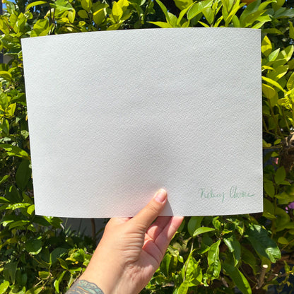 A hand is holding a blank white sheet of paper against a backdrop of green leaves and foliage.