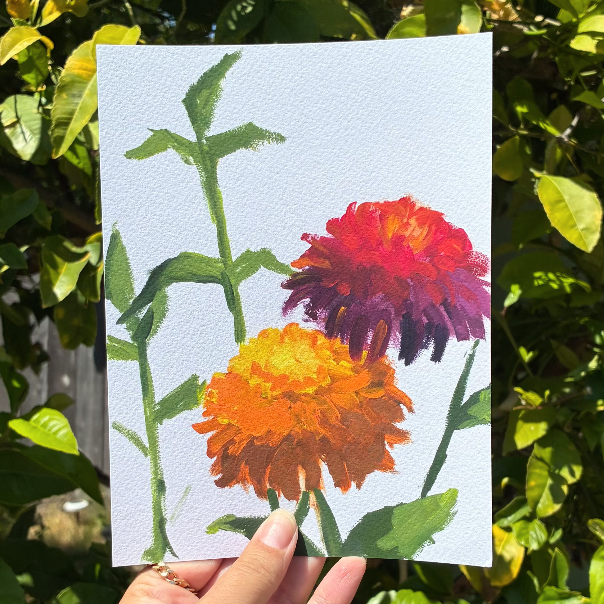 A hand is holding a piece of paper with a painting of two colorful flowers, one orange and one purple, against a background of green leaves.