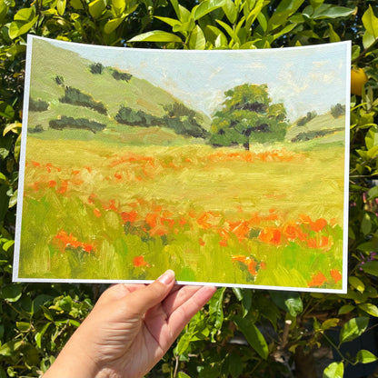 A hand is holding up a painting of a field with trees and flowers, against a backdrop of green foliage.