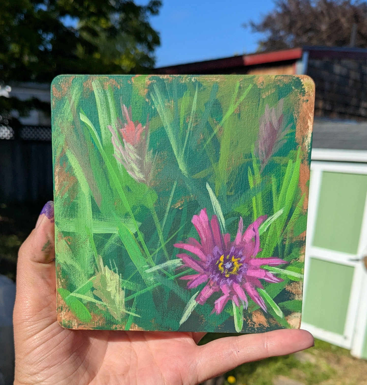A hand is holding a square piece of art featuring a vibrant pink flower surrounded by lush green foliage.