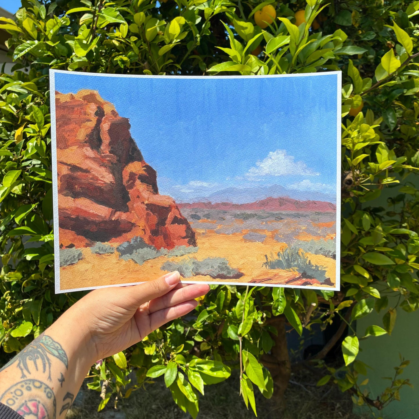 A hand holding a framed painting of a desert landscape with a rocky cliff and a blue sky.
