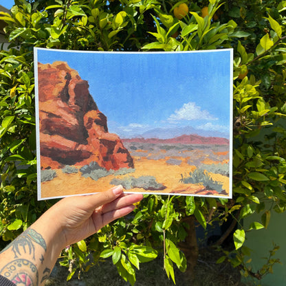 A hand holding a framed painting of a desert landscape with a rocky cliff and a blue sky.